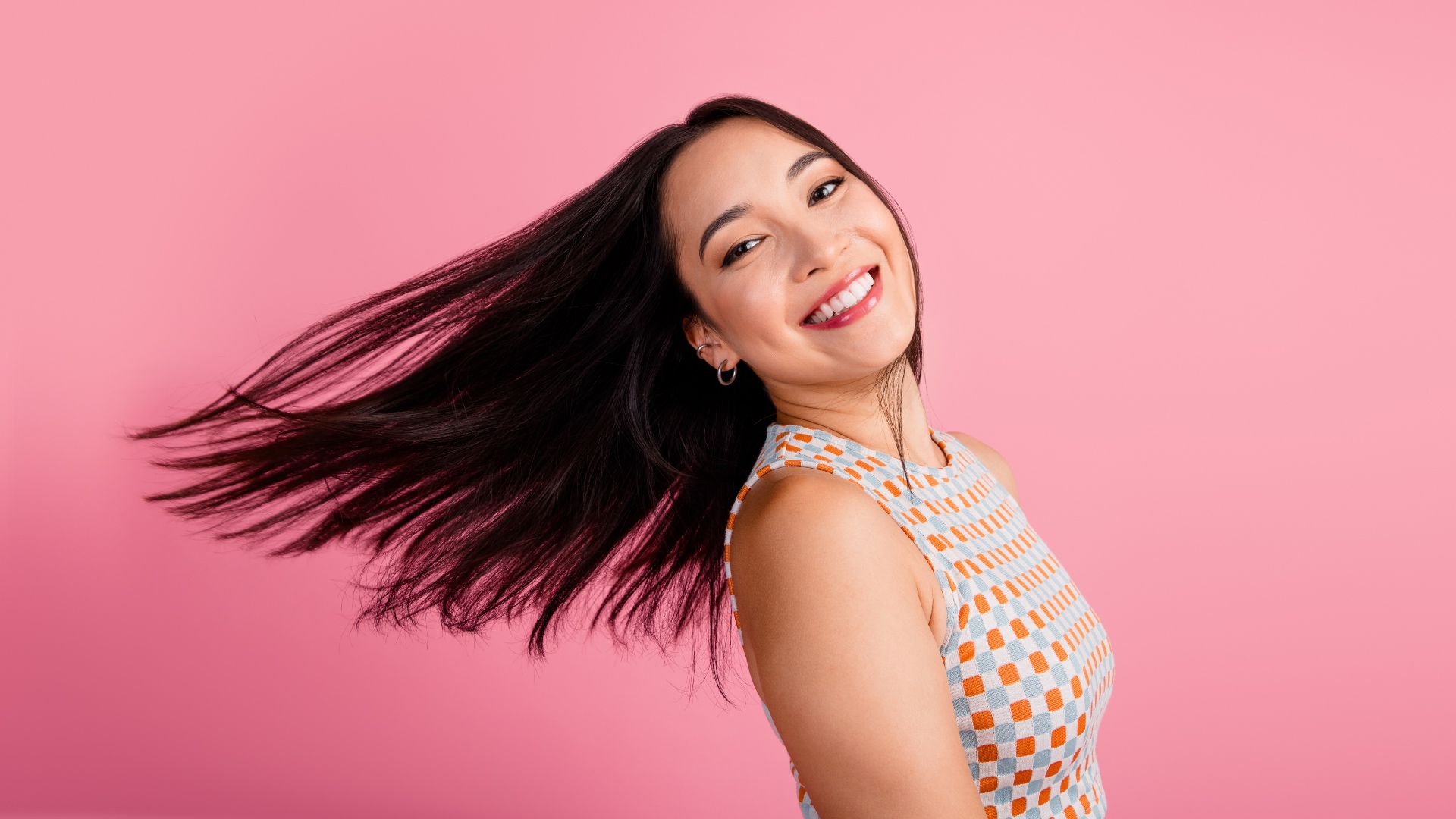 Joyful young woman with vibrant brunette hair flipping in front of a pink background, showcasing trendy summer fashion and lifestyle